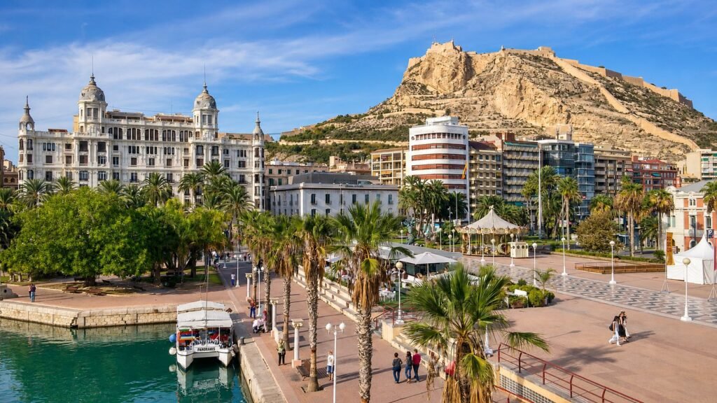 Panoramic view of Alicante city port and promenade with Santa Bárbara Castle on the hill under a clear blue sky.