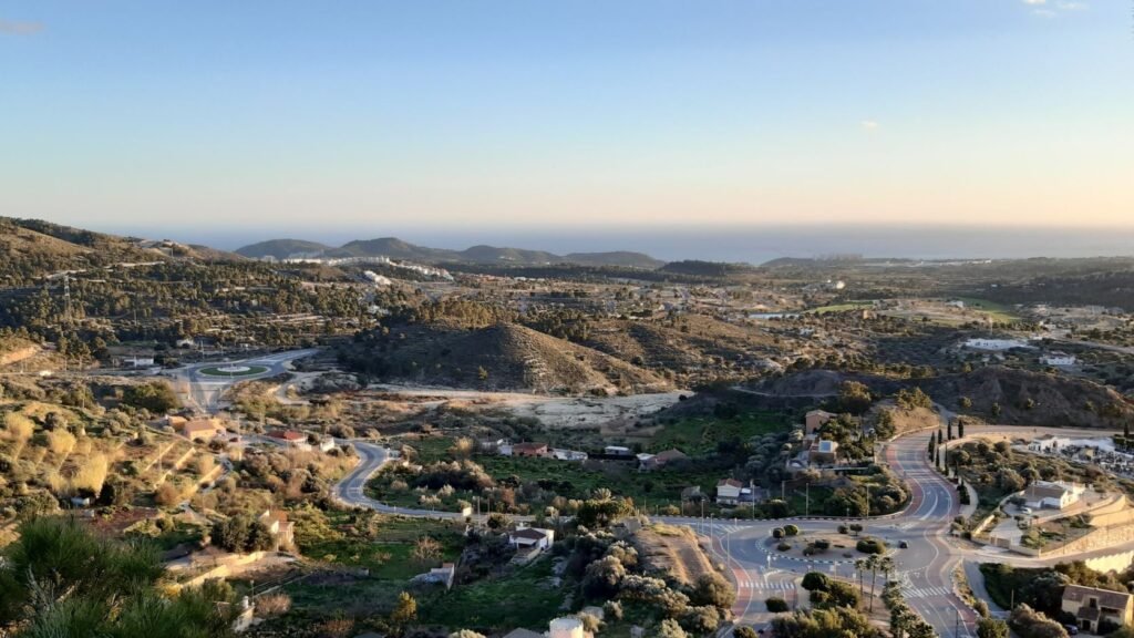 Elevated panoramic view of Xixona countryside, winding roads, and pine-covered hills extending towards the distant Mediterranean sea.